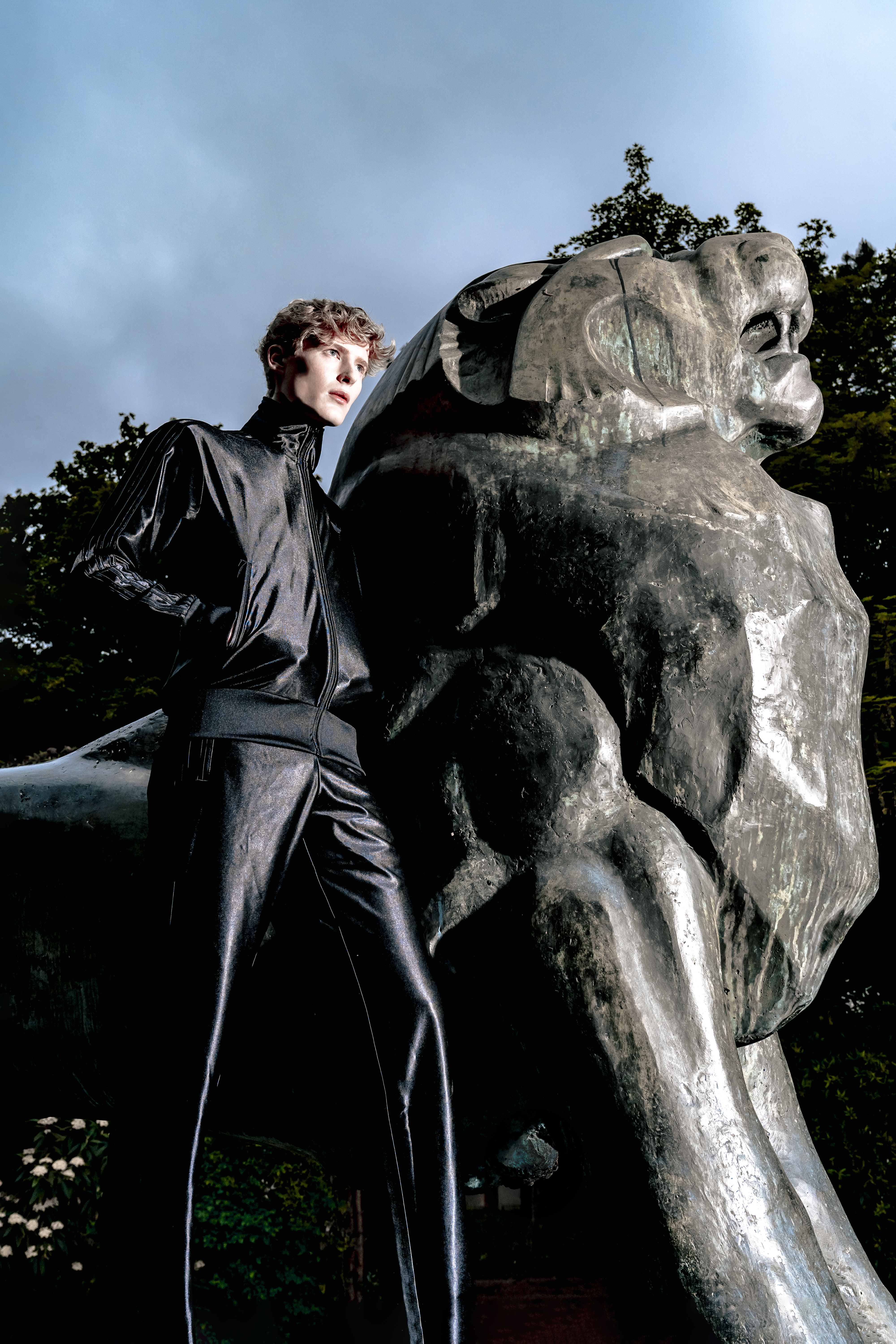 Model wearing a metallic black tracksuit standing beside a lion statue.