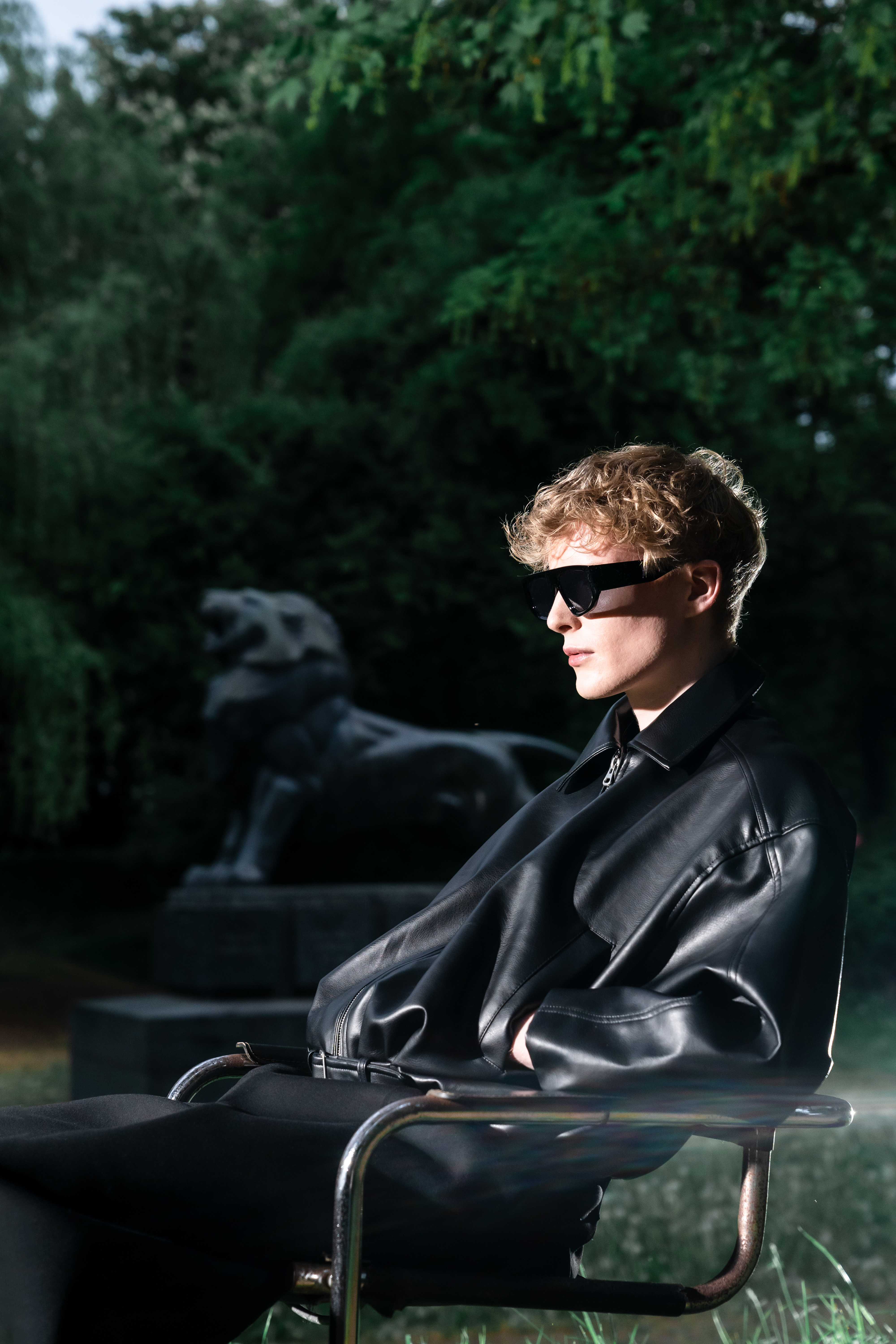 Model wearing an oversized leather jacket, dark sunglasses and relaxed trousers beside the lion statue.