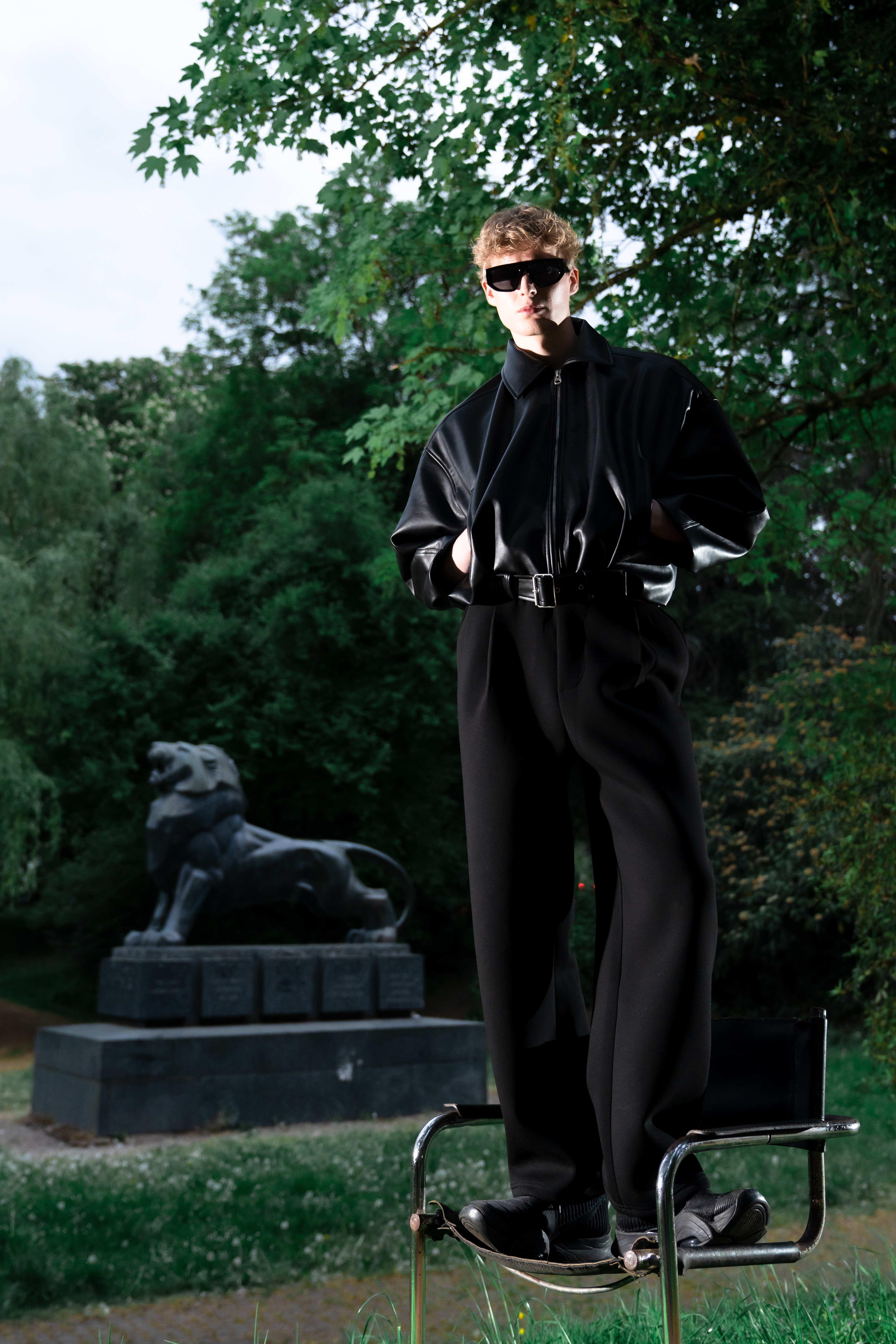 Model standing on a chair wearing an oversized leather jacket, dark sunglasses and relaxed trousers.