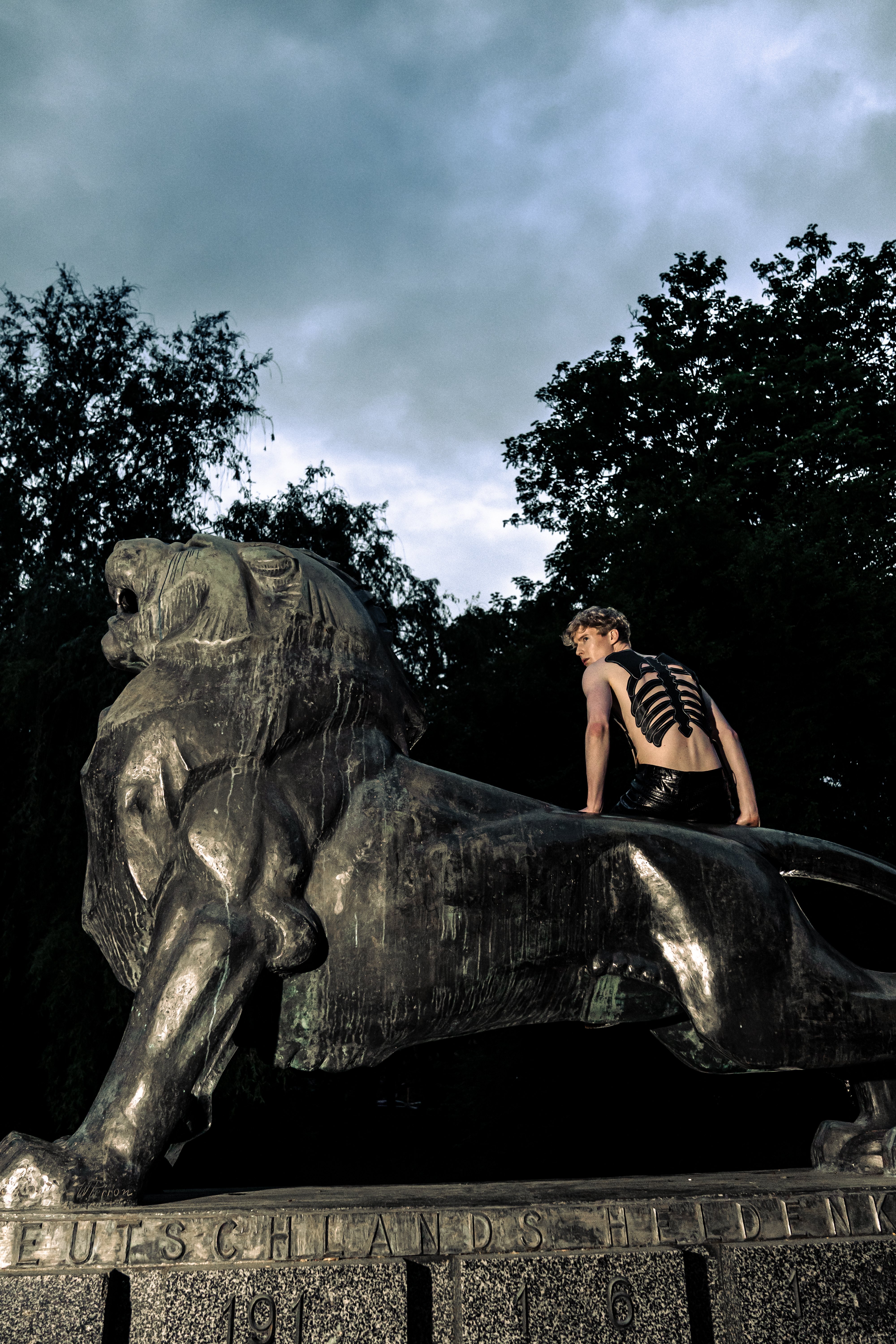 Model wearing a black skeleton cuirass and snakeskin leather trousers seated on the lion statue.