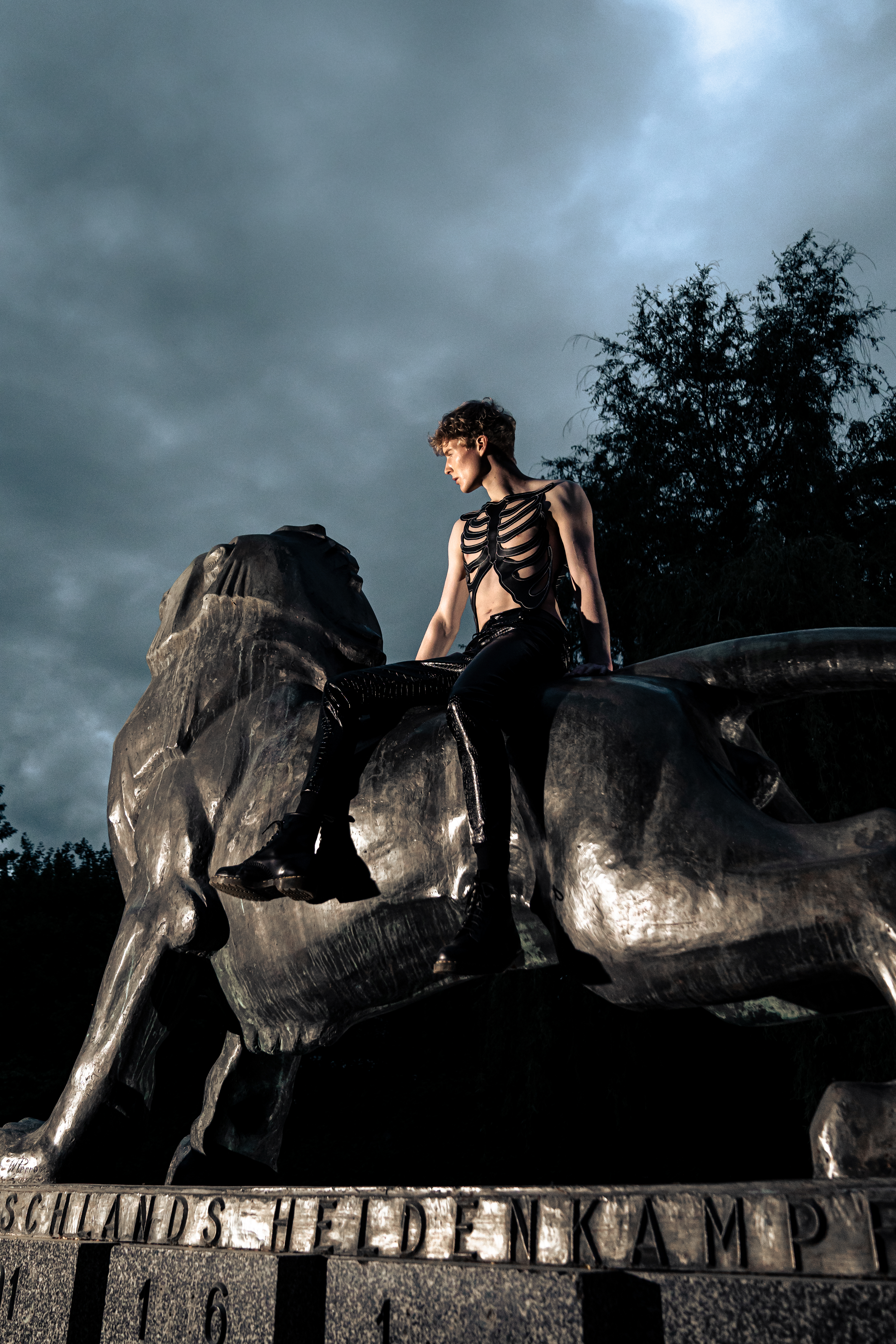 Model wearing a black skeleton cuirass and snakeskin leather trousers seated on the lion statue, looking towards the horizon.