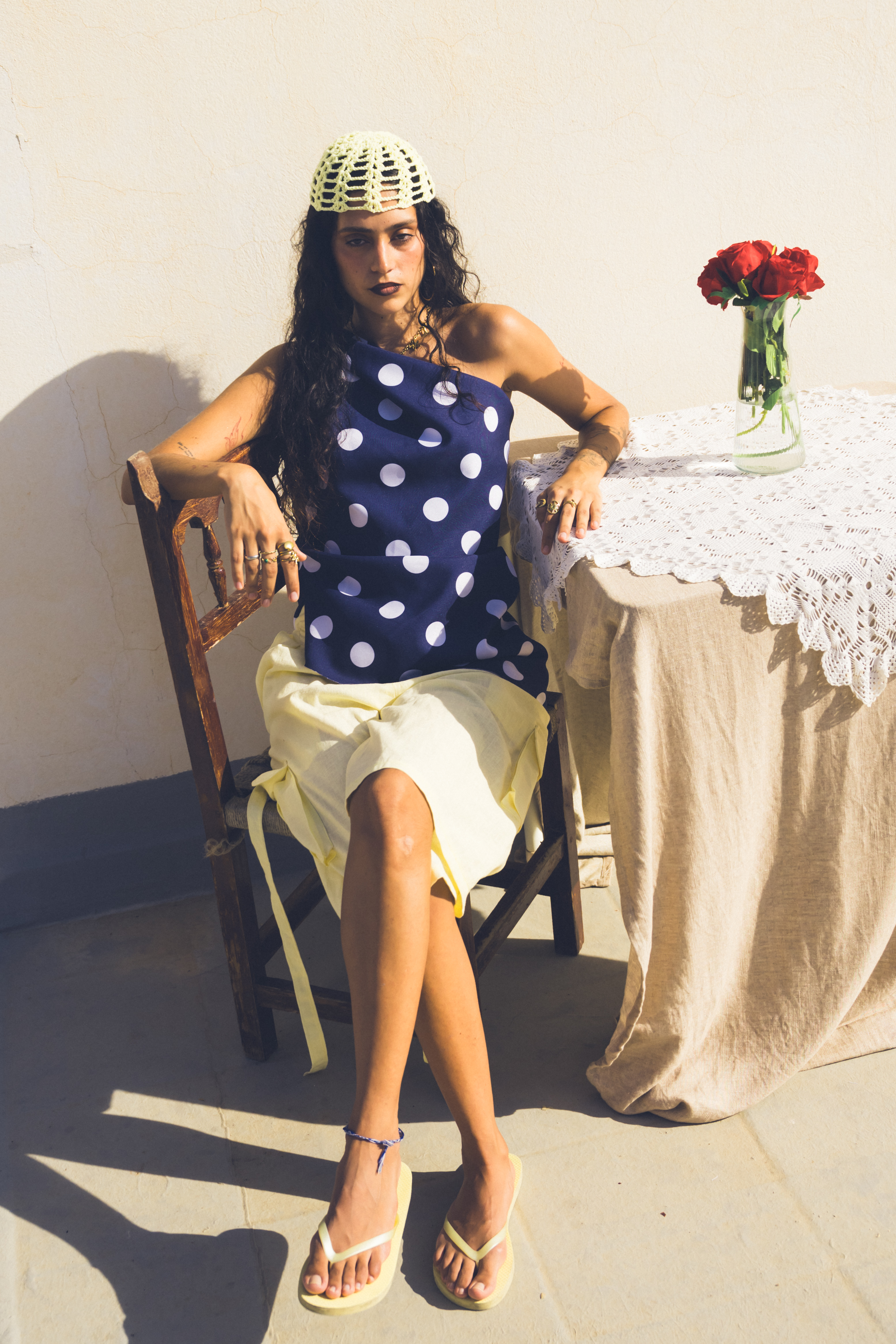 Modelo sentada junto a una mesa con un pequeño florero con una rosa rosa en un entorno rural.