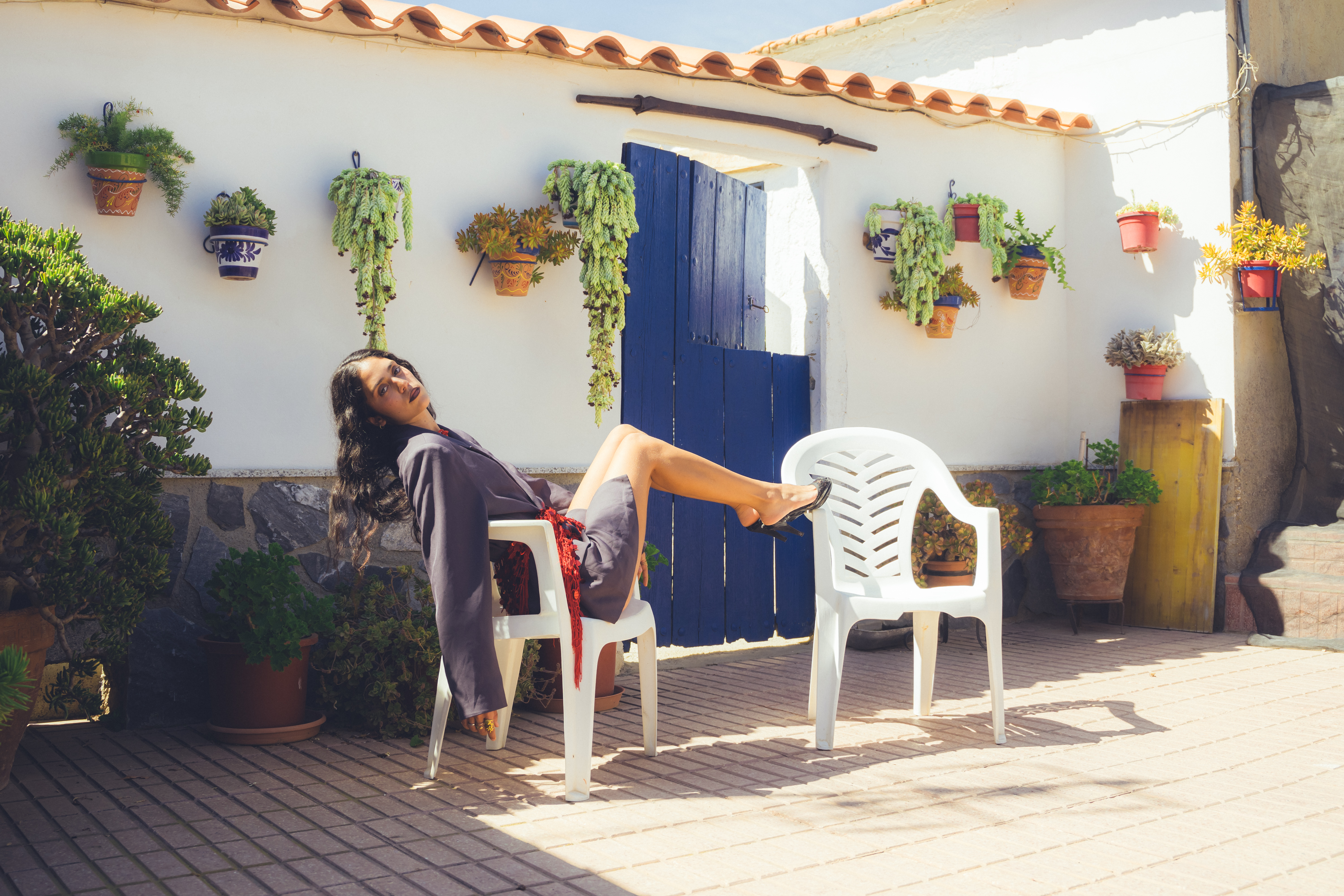 Modelo sentada frente a la entrada de una casa, con las piernas apoyadas sobre otra silla, en una actitud relajada.