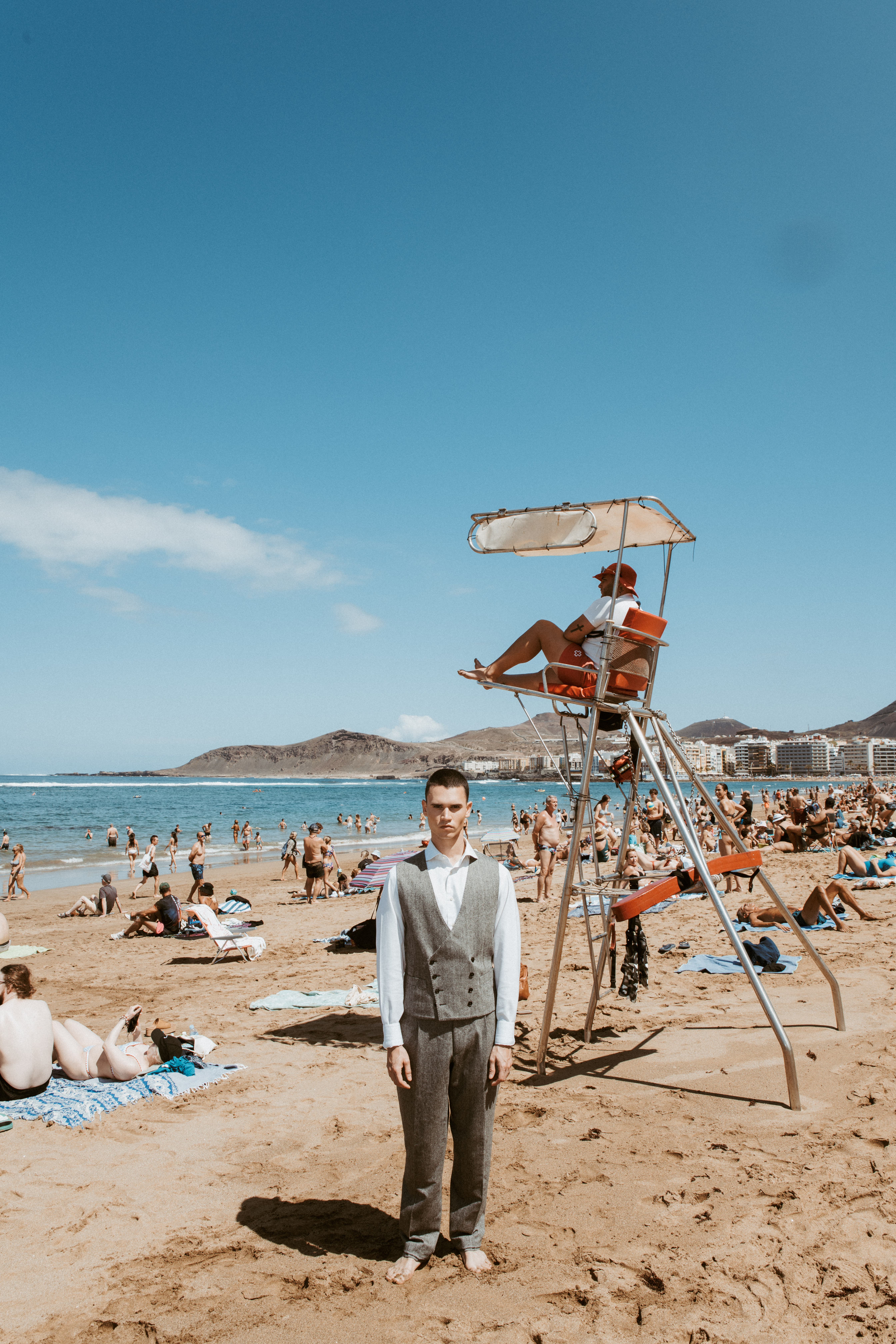 Modelo descalzo posando entre turistas con estilismo de pantalón de vestir y chaqueta en un editorial de verano
