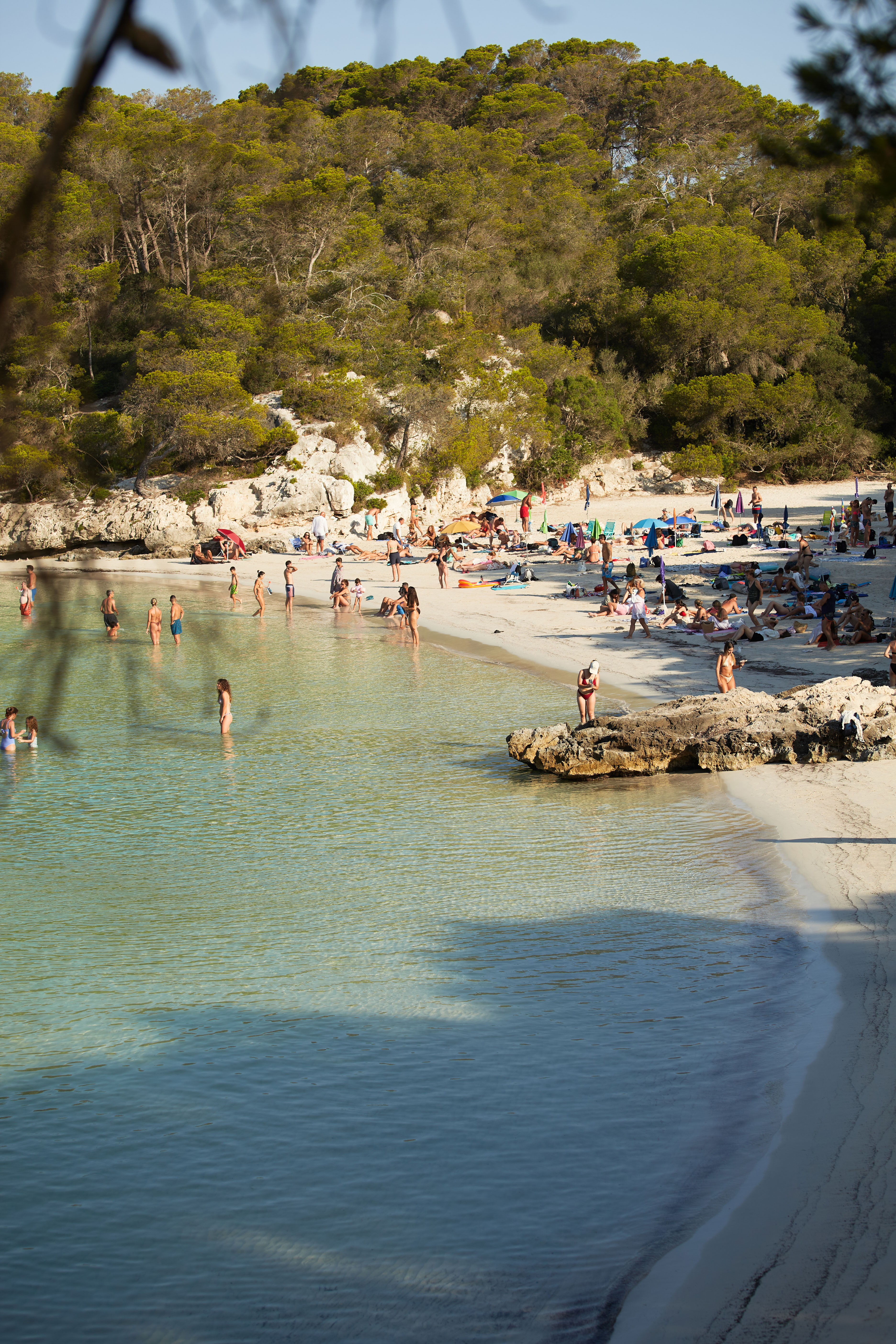 Veraneantes en Cala Turqueta, Menorca, junto a un mar turquesa rodeado de pinos mediterráneos