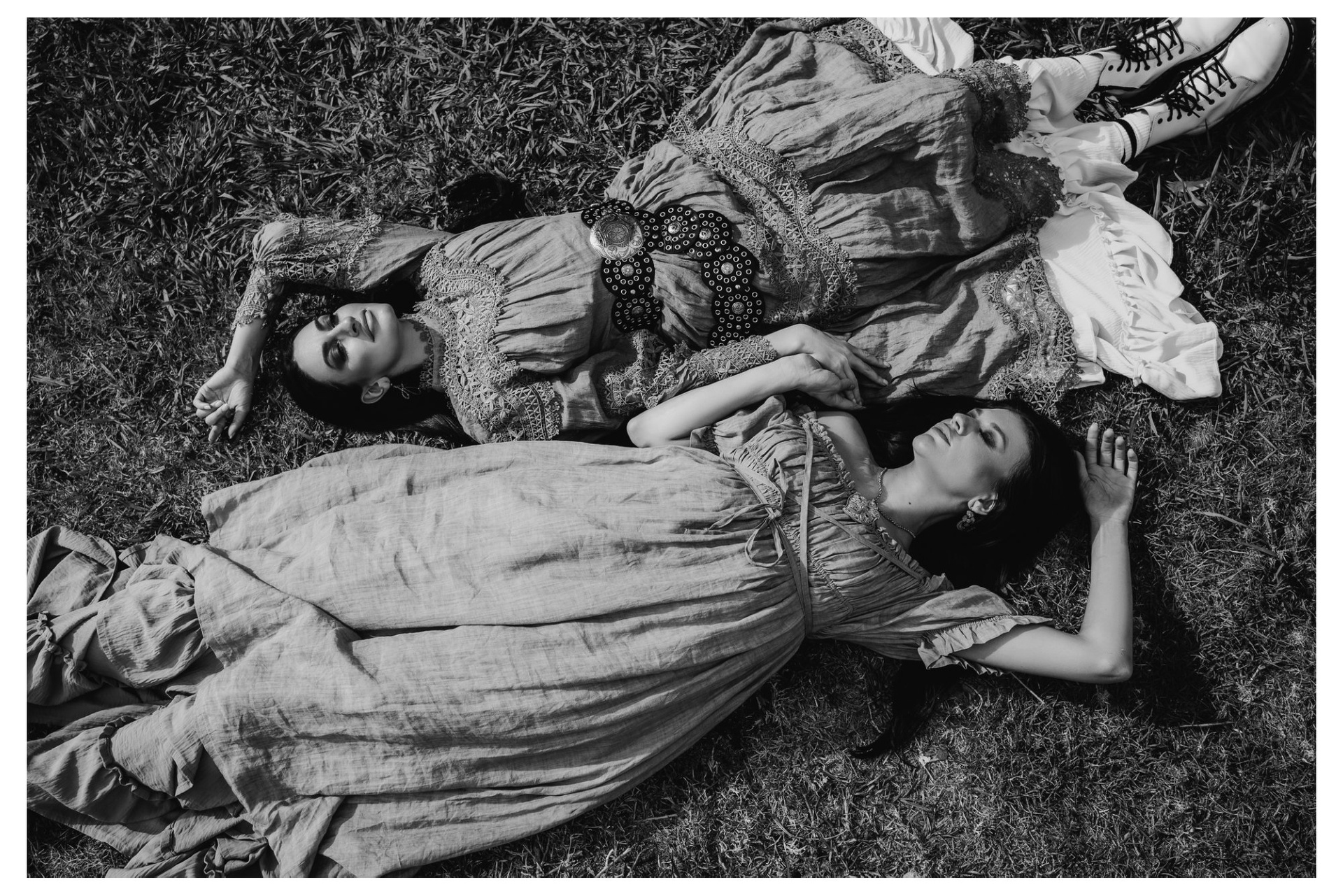 Two women lying on the grass wearing ruffled dresses, layered petticoats, beaded bonnets, sports socks, chunky boots and bold belts